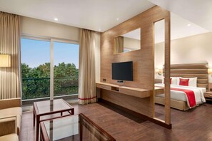 Quarto de hotel com uma janela grande, uma TV de tela plana, uma cama, mesa de cabeceira e área de estar com uma mesa e cadeiras.