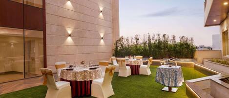 An outdoor dining area with tables and chairs, a building with a modern design, and a clear sky.