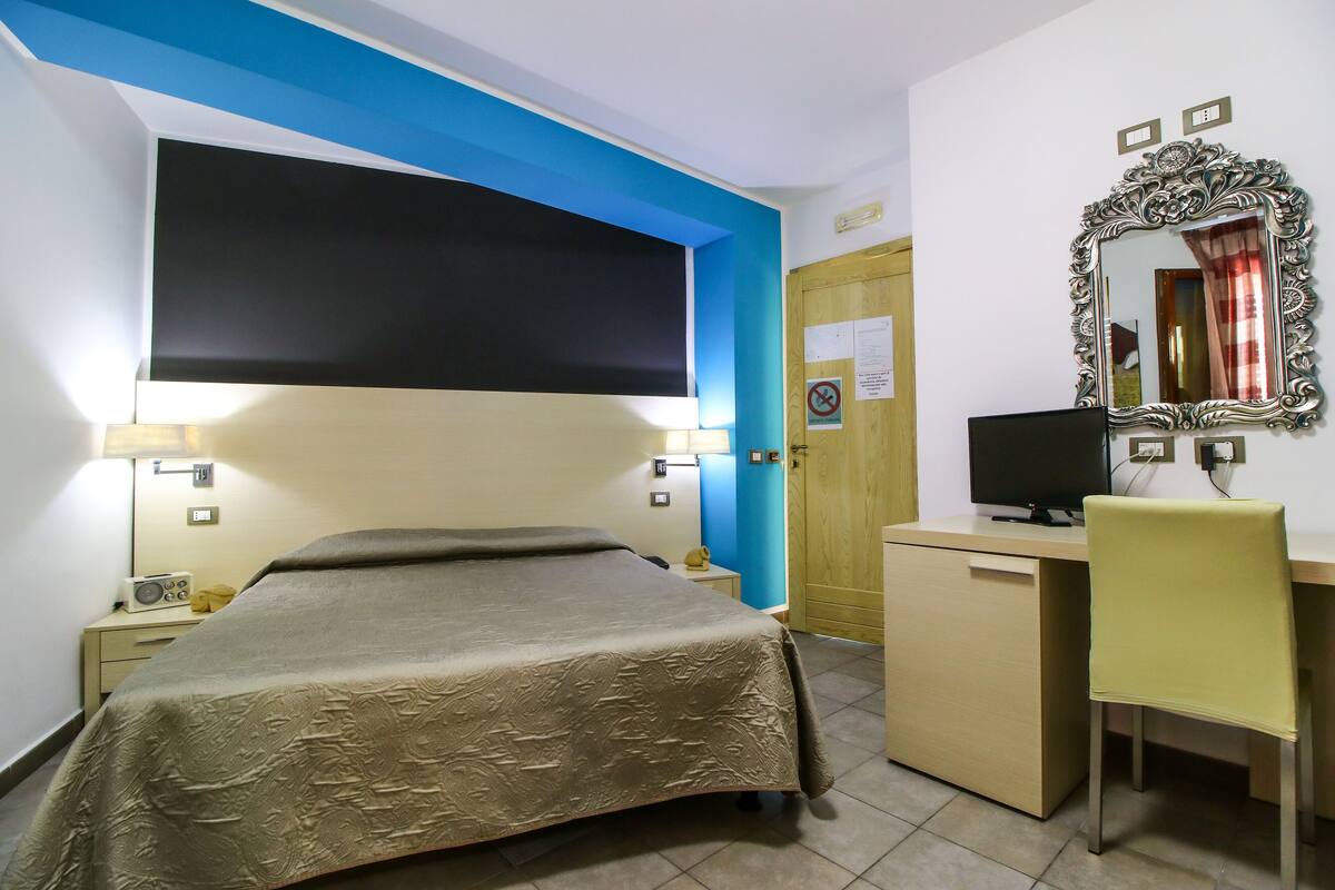 A hotel room with a bed, desk, chair, and a large mirror.
