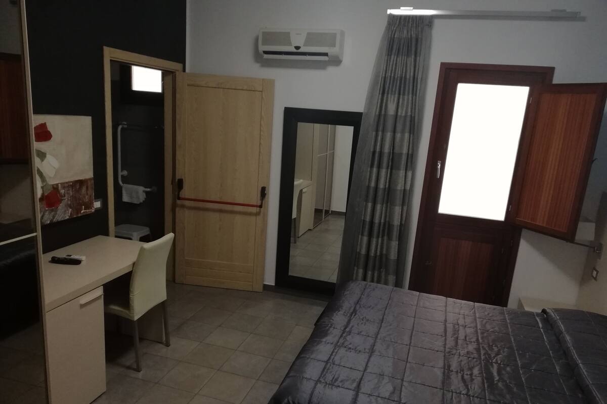 A bedroom with a bed, a desk, a chair, a wardrobe, a mirror, and a window with curtains.