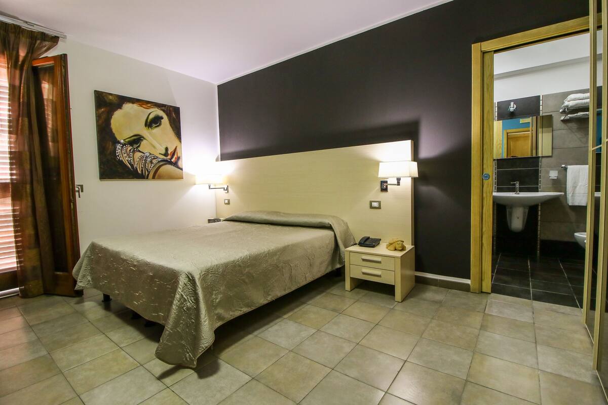 A hotel room with a bed, bedside table, lamp, and a painting on the wall.