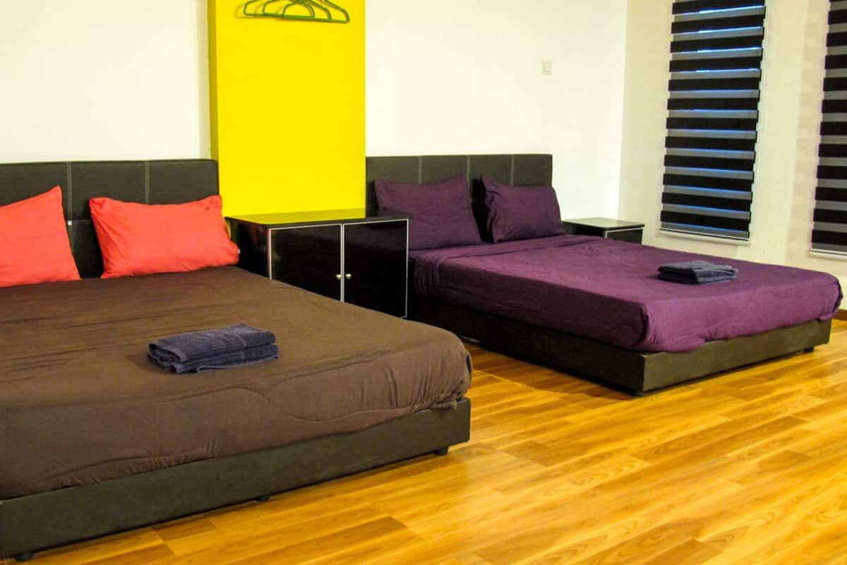 A hotel room with two beds, wooden flooring, a yellow wall decoration, and a window with blinds.