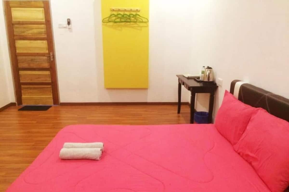 A room with a bed, a small table, a chair, a yellow wall art, and a wooden door.