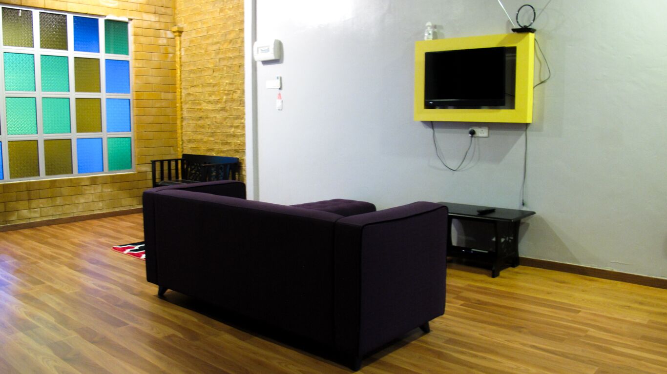 A room with a purple sofa, a small black table, a TV mounted on a yellow frame, and a window with colorful glass panes.