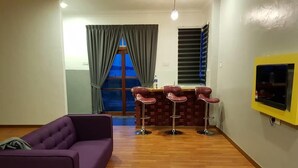 A living room with a purple sofa, a small ottoman, and a bar area with stools. There is a TV mounted on the wall and a window with curtains. - Multaqa Suite & Residence (Marang)