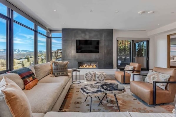 A modern living room with a fireplace, large windows, and a view of the mountains. - One Snowmass West 303 (SNOWMASS VILLAGE)