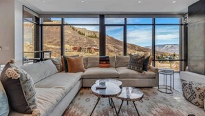 A modern living room with a large sectional sofa, two round coffee tables, and a view of a mountainous landscape through floor-to-ceiling windows. - One Snowmass West 303 (SNOWMASS VILLAGE)