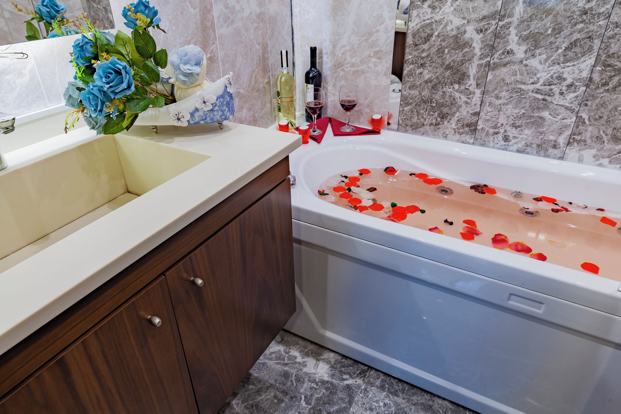 A bathroom with a bathtub filled with rose petals, a sink with a vase of blue flowers, and two bottles of wine on the counter.
