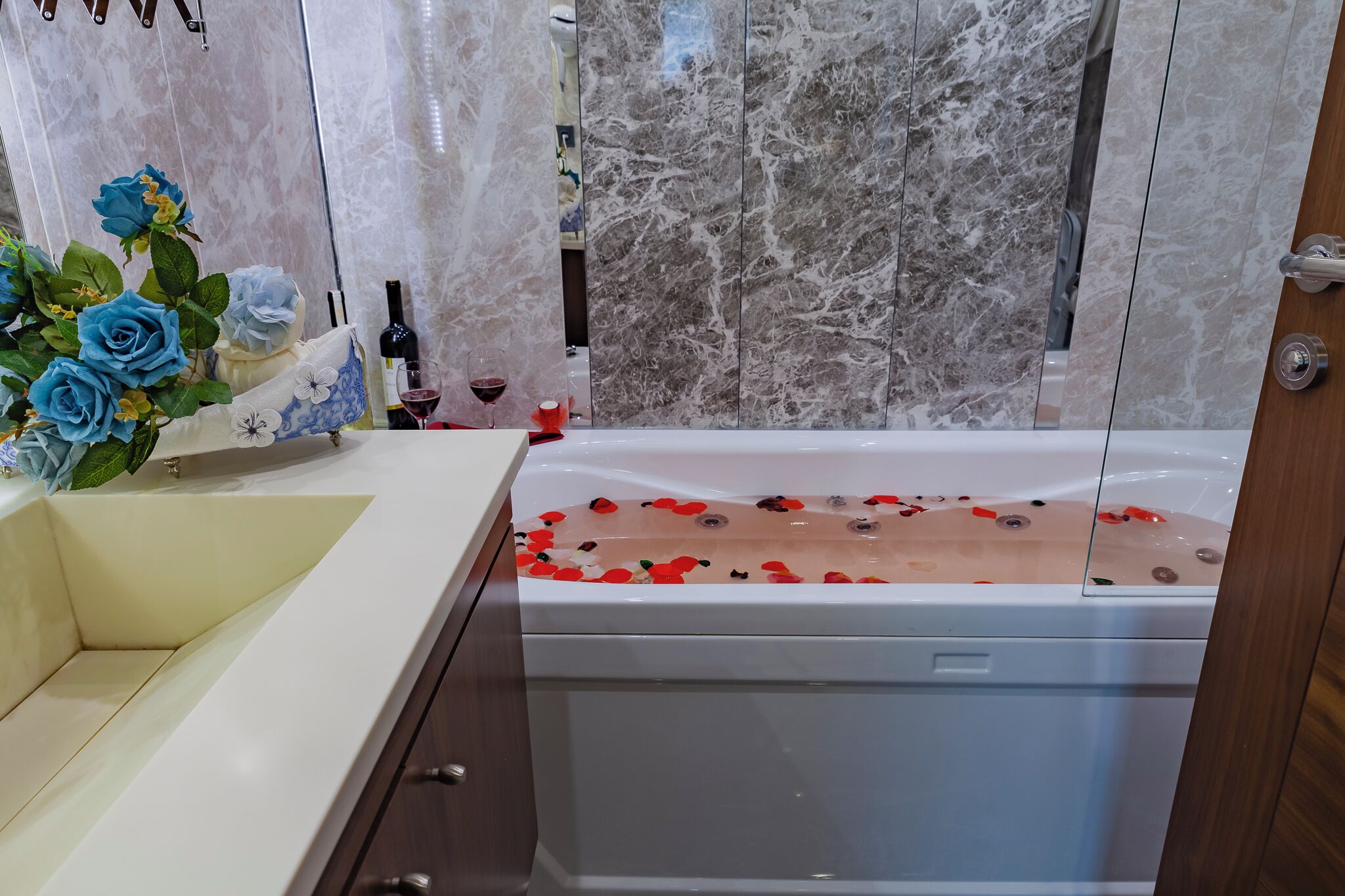 A bathroom with a marble wall, a bathtub with rose petals, and a vase of blue roses.