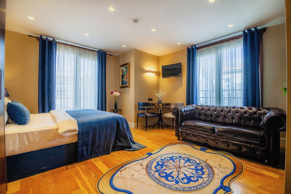 A hotel room with a bed, a leather sofa, a dining table with chairs, a TV mounted on the wall, and a patterned rug.
