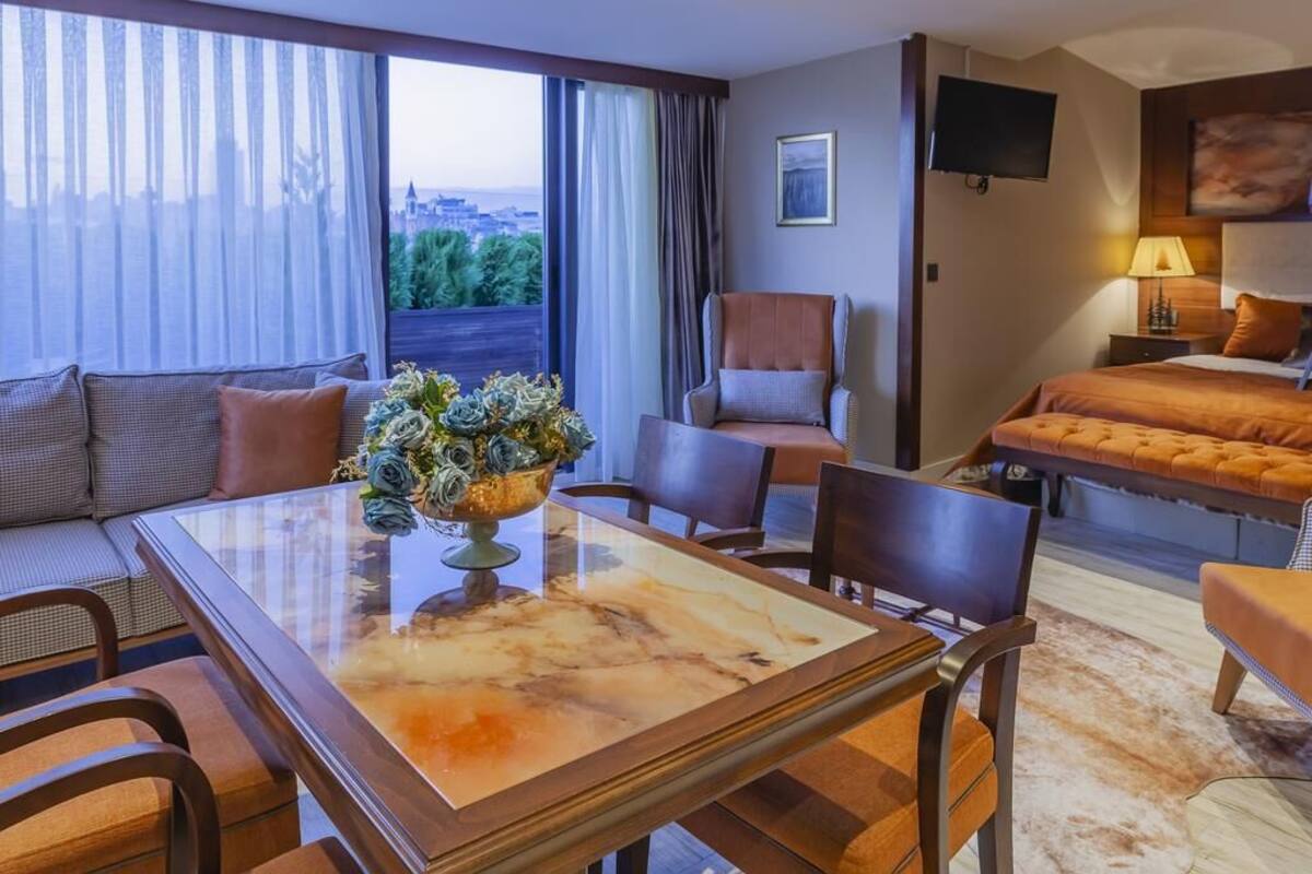 A hotel room with a large window, a sofa, a dining table, a TV, and a bed.