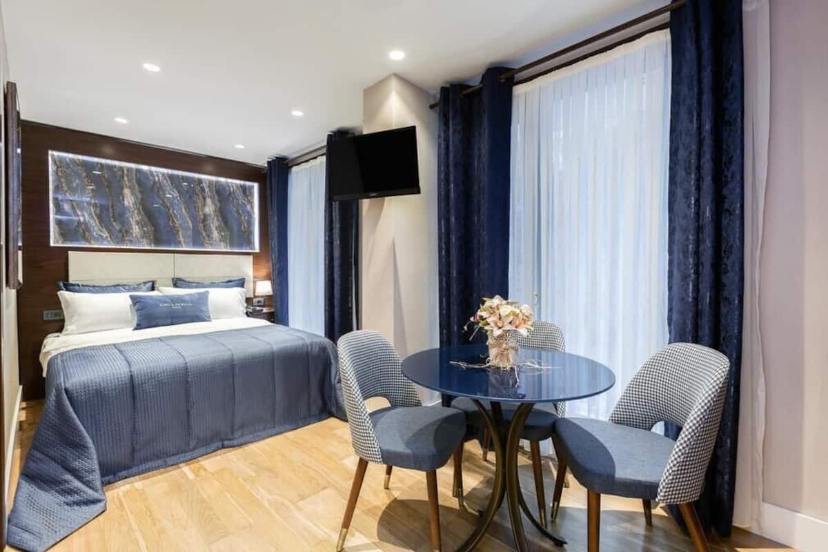 A modern hotel room with a large bed, a round table with chairs, a flat-screen TV, and a large window with curtains.
