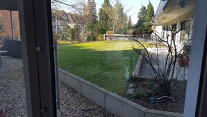 A view through a window of a residential area with a well-maintained lawn, a concrete retaining wall, and a house with a visible garage door. - Cozy luxury 4 bedroom. In the heart of Heidelberg (Kirchheim)