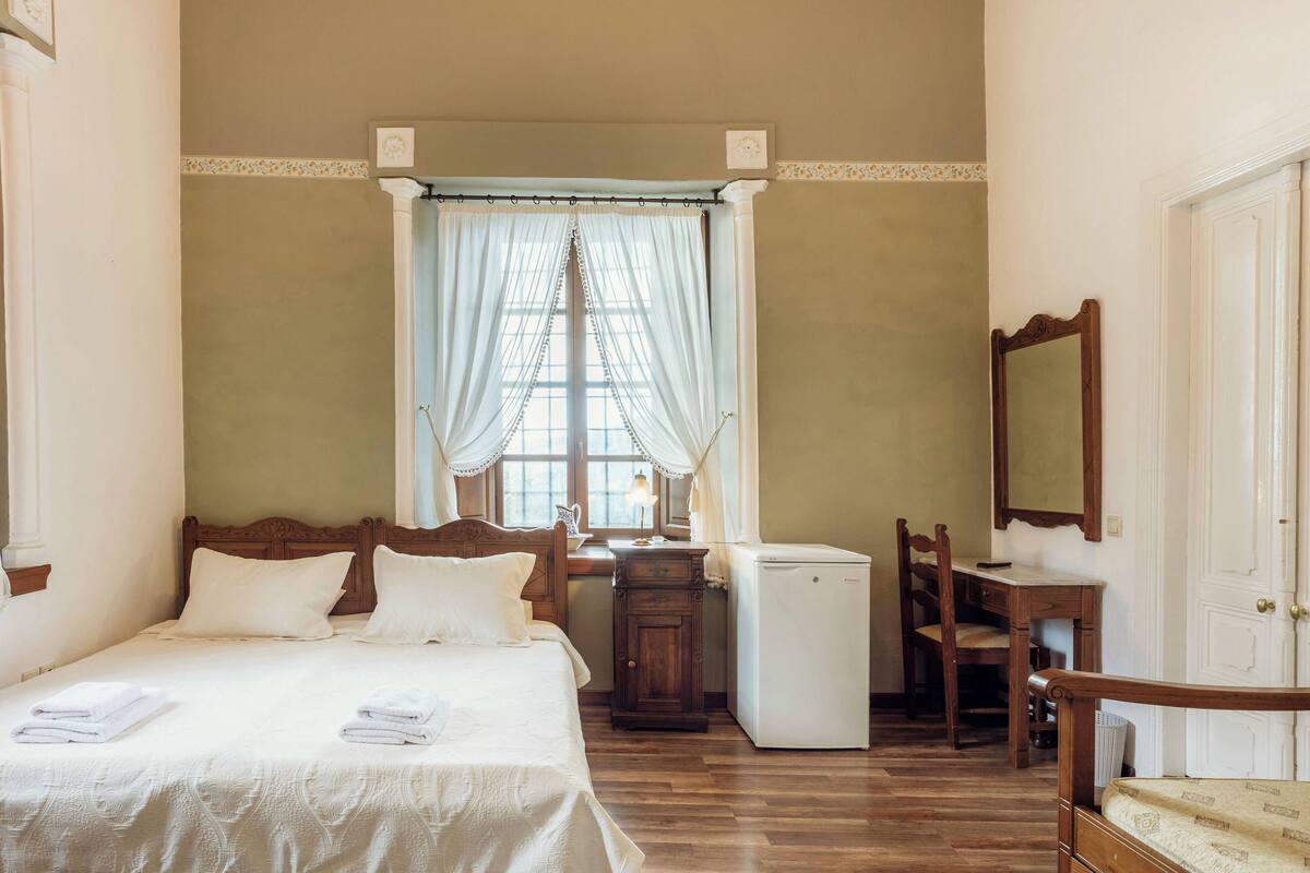 Un dormitorio con cama, escritorio, silla, espejo y un refrigerador pequeño.