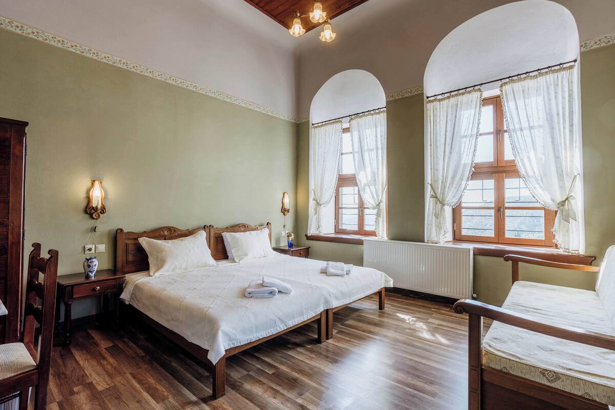 Un dormitorio con una cama grande, dos mesitas de noche, una silla y un ventanal con cortinas.