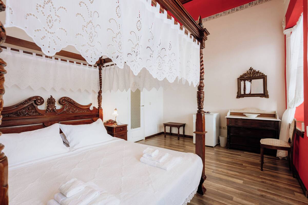 Una cama con dosel, ropa de cama blanca y dosel de encaje, una mesita de noche de madera con lámpara, un aparador de madera y una silla en una habitación con suelo de madera y paredes rojas.