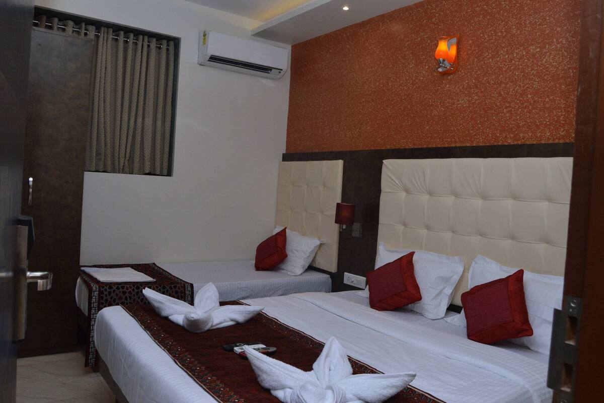 Quarto de hotel com duas camas, cada uma com lençóis brancos e almofadas vermelhas. Há uma pequena mesa com uma toalha e um controle remoto. O quarto possui uma parede em tom de marrom e uma janela com cortinas.