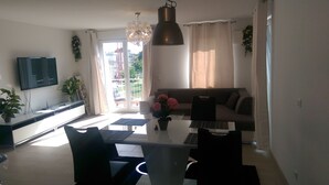 Ein modernes Wohnzimmer mit Flachbildfernseher, einem Esstisch mit Stühlen, einer Couch und einem großen Fenster mit Vorhängen.