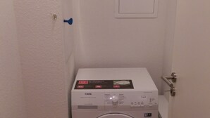 Une petite machine à laver blanche, installée dans un petit espace buanderie fermé, avec une porte et une prise de courant au sol.