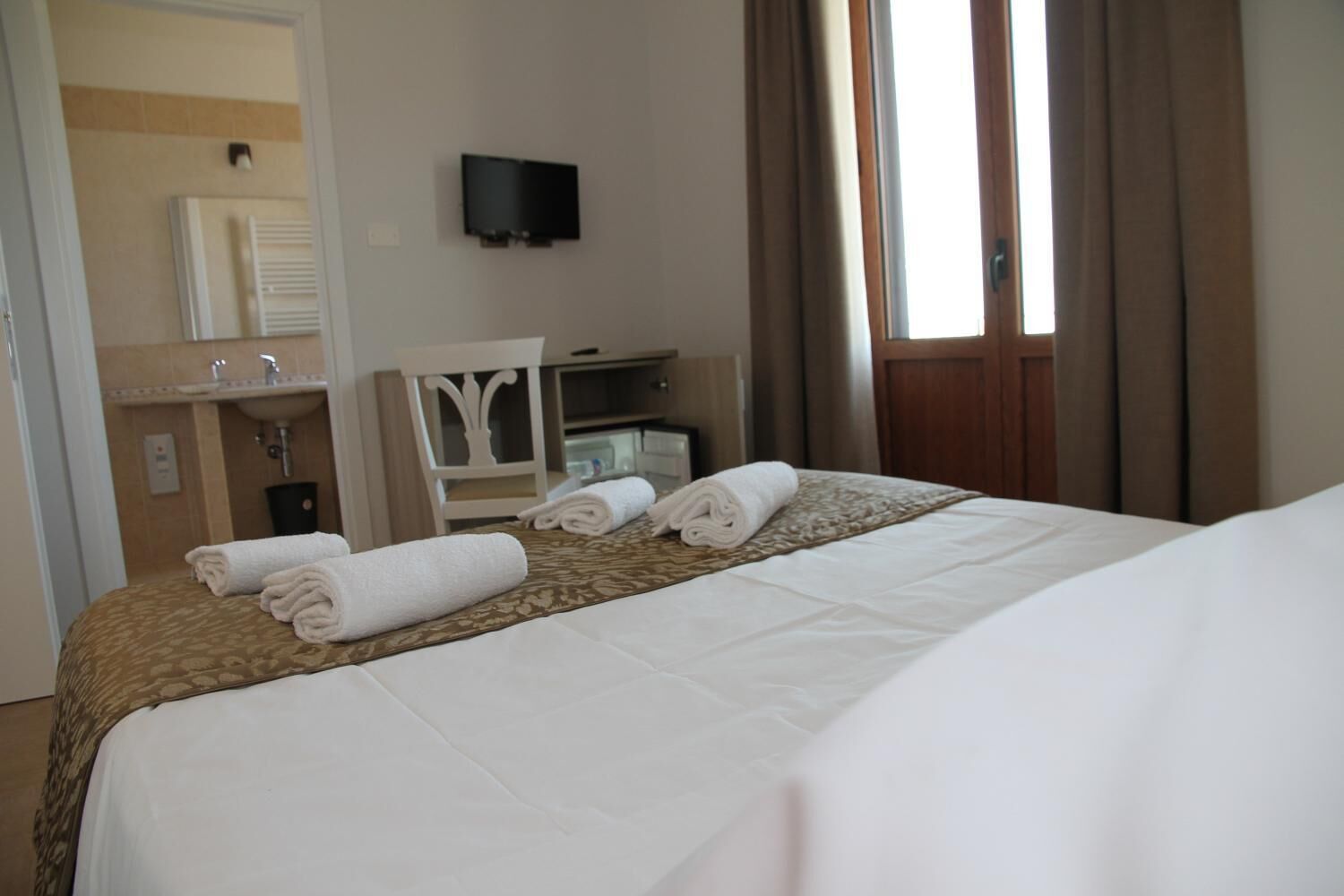 Habitación de hotel con cama, televisión montada en la pared, baño con lavabo y espejo, y ventana con cortinas.