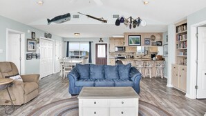 Amplia sala de estar con un sofá azul, una mesa de centro blanca y un tapete estampado. Se observa una zona de cocina con armarios de madera y una mesa de comedor con sillas.
