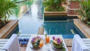 Escena junto a la piscina con una mesa preparada para dos, que incluye una bebida y un cuenco de frutas, rodeada de sillones de descanso y vistas a la piscina.