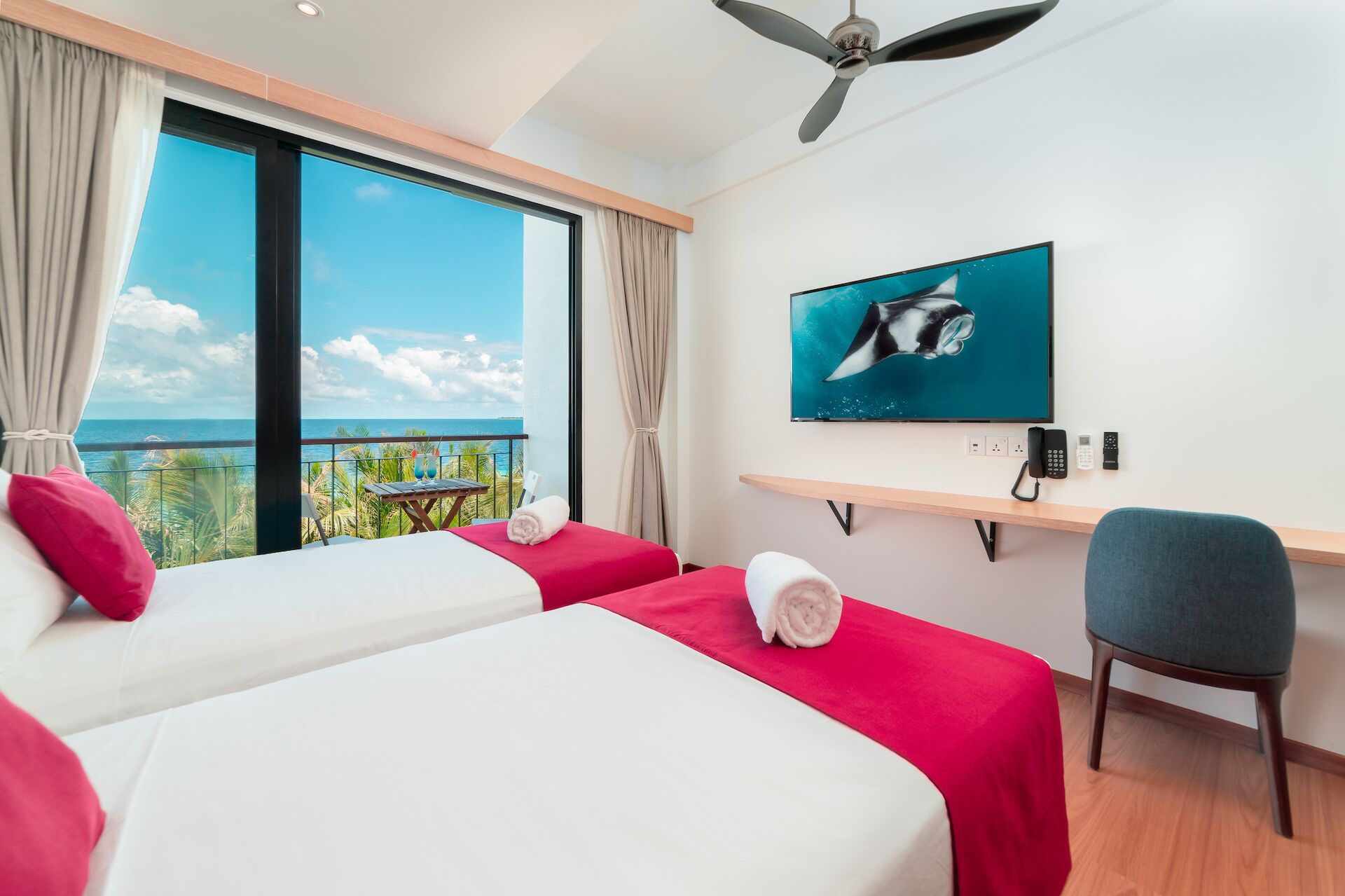 Ein Hotelzimmer mit zwei Betten, einem Schreibtisch, einem Stuhl, einem Fernseher und einem Balkon mit Meerblick.