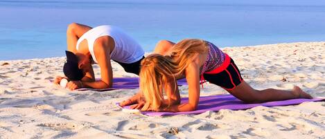 Two people doing yoga on purple mats on a sandy beach with the ocean in the background.
