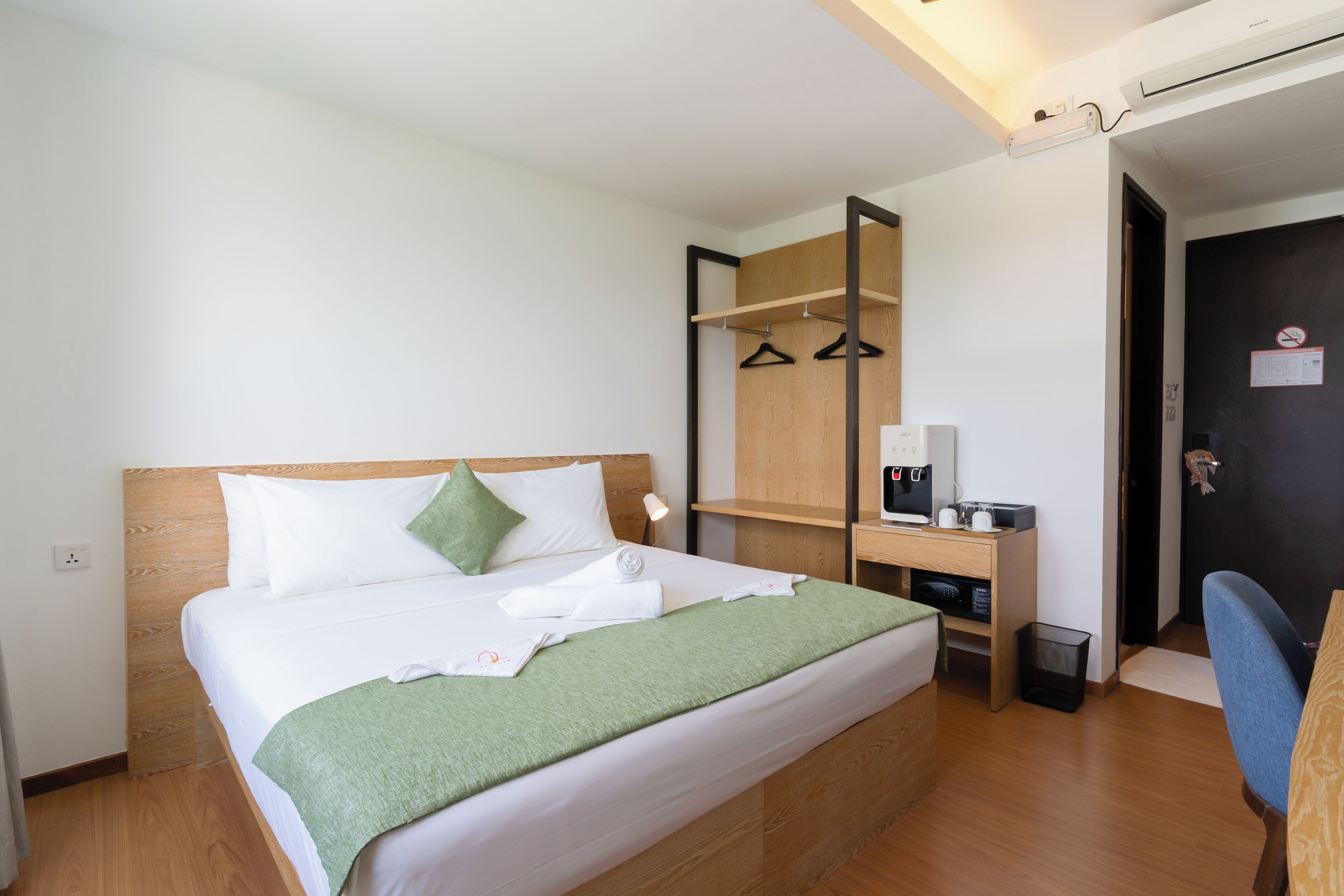 A neatly made bed with a green blanket, a bedside table with a coffee maker, and a wooden headboard.