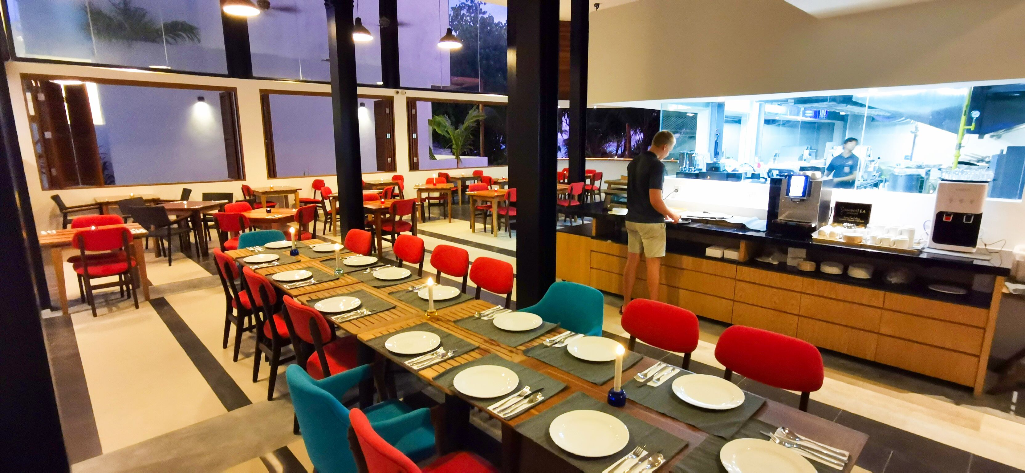 Een restaurant met meerdere eettafels, rode en blauwe stoelen, en een zichtbare keukenruimte achter een glazen scheidingswand.