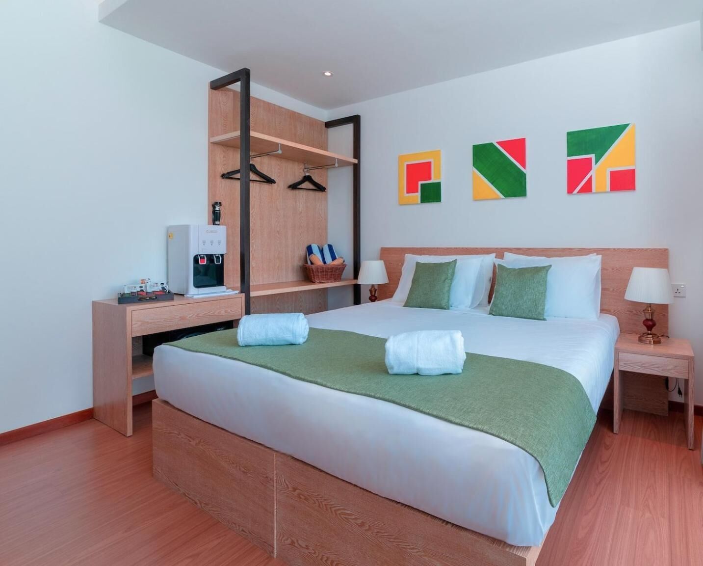 A hotel room with a bed, a desk with a coffee machine, a wardrobe, and a wall with colorful geometric art.