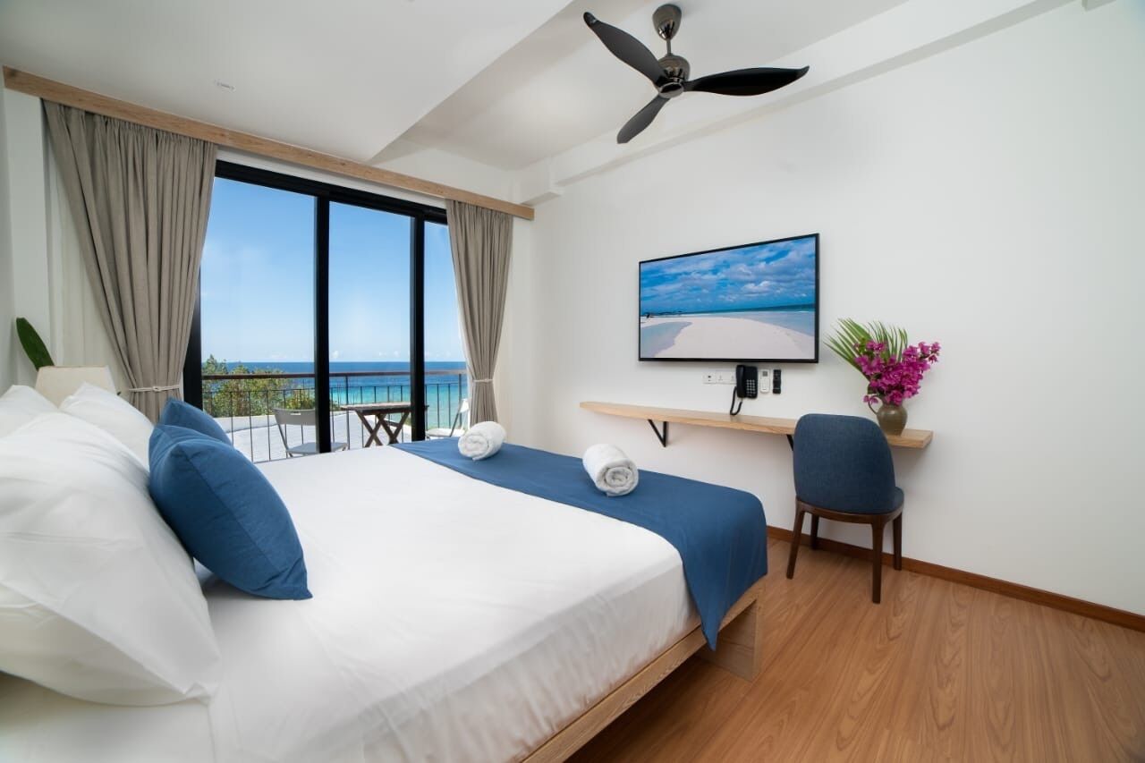 A hotel room with a bed, a desk, a chair, a TV, and a view of the sea.