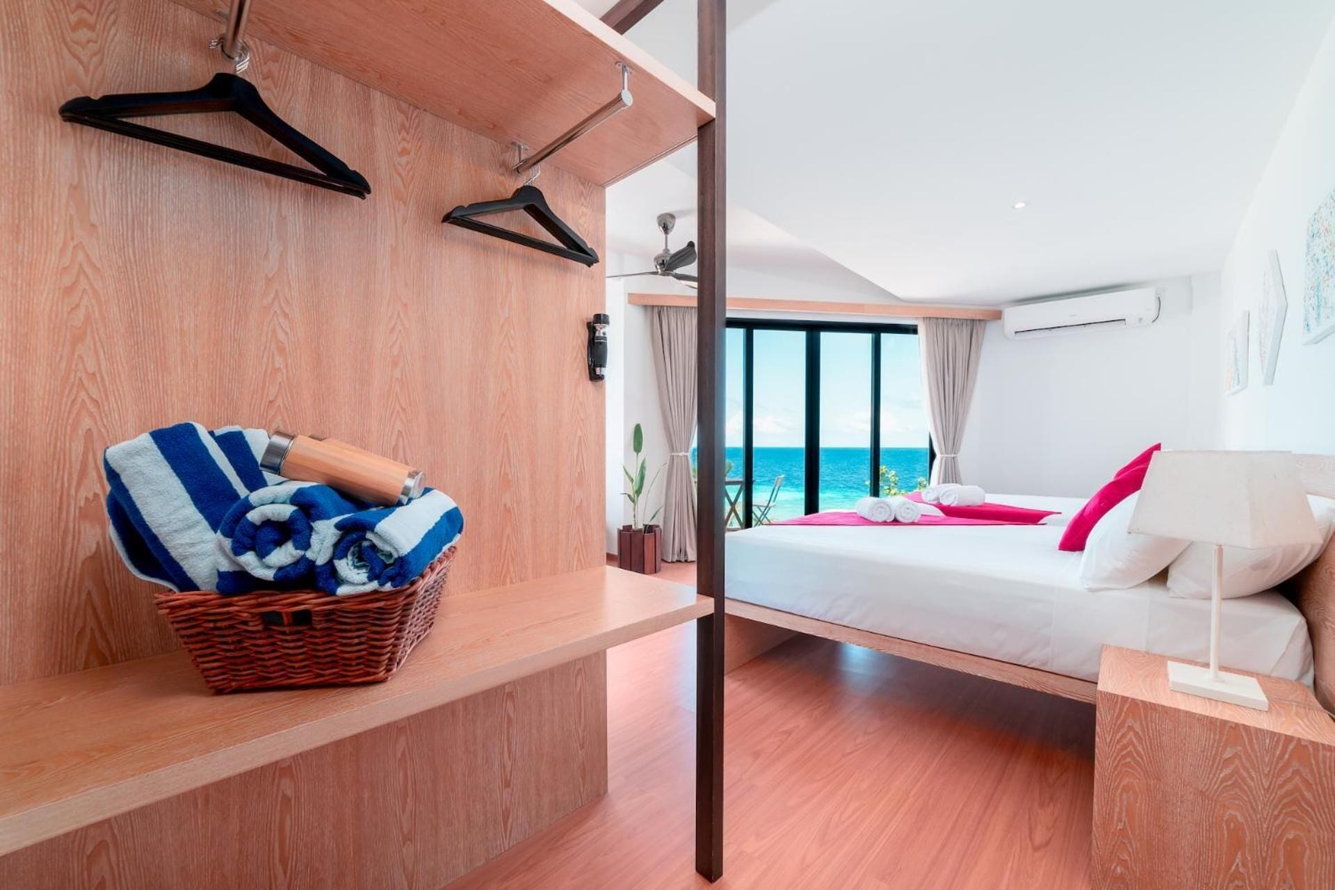Een moderne hotelkamer met een houten vloer, een bed met witte en roze beddengoed, een rieten mand met handdoeken en uitzicht op de oceaan door grote ramen.