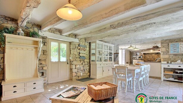 Une cuisine rustique aux murs de pierre, avec des poutres en bois et un coin repas doté de chaises blanches.