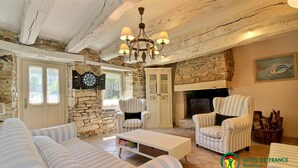 Un salon chaleureux avec des canapés à rayures, une table basse blanche et une cheminée en pierre. On y trouve deux fauteuils et un meuble avec des portes en verre. Un lustre est suspendu au plafond, et un jeu de fléchettes est accroché au mur.