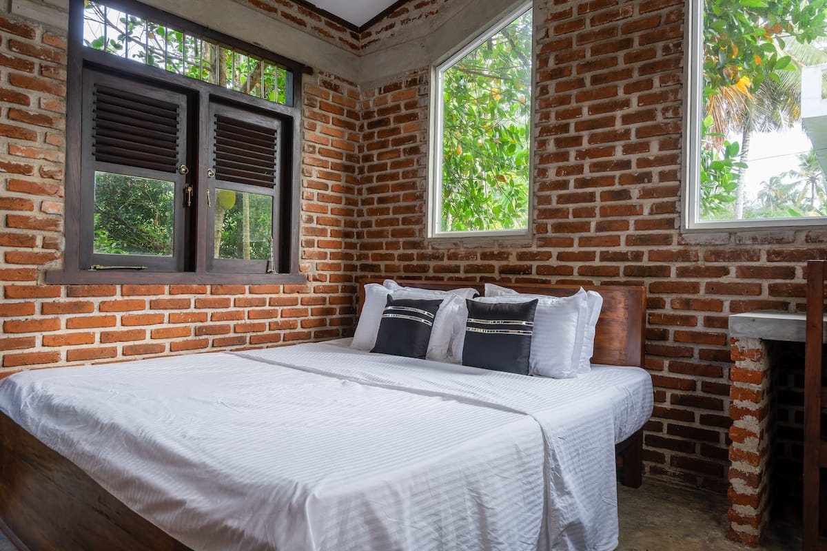 A room with a brick wall, two windows with shutters, a bed with white bedding, and a nightstand.
