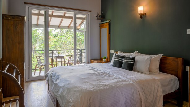 A bedroom with a bed, a nightstand, a mirror, and a sliding glass door leading to an outdoor area with a table and chairs. - Dollyz Home (Madampagama)