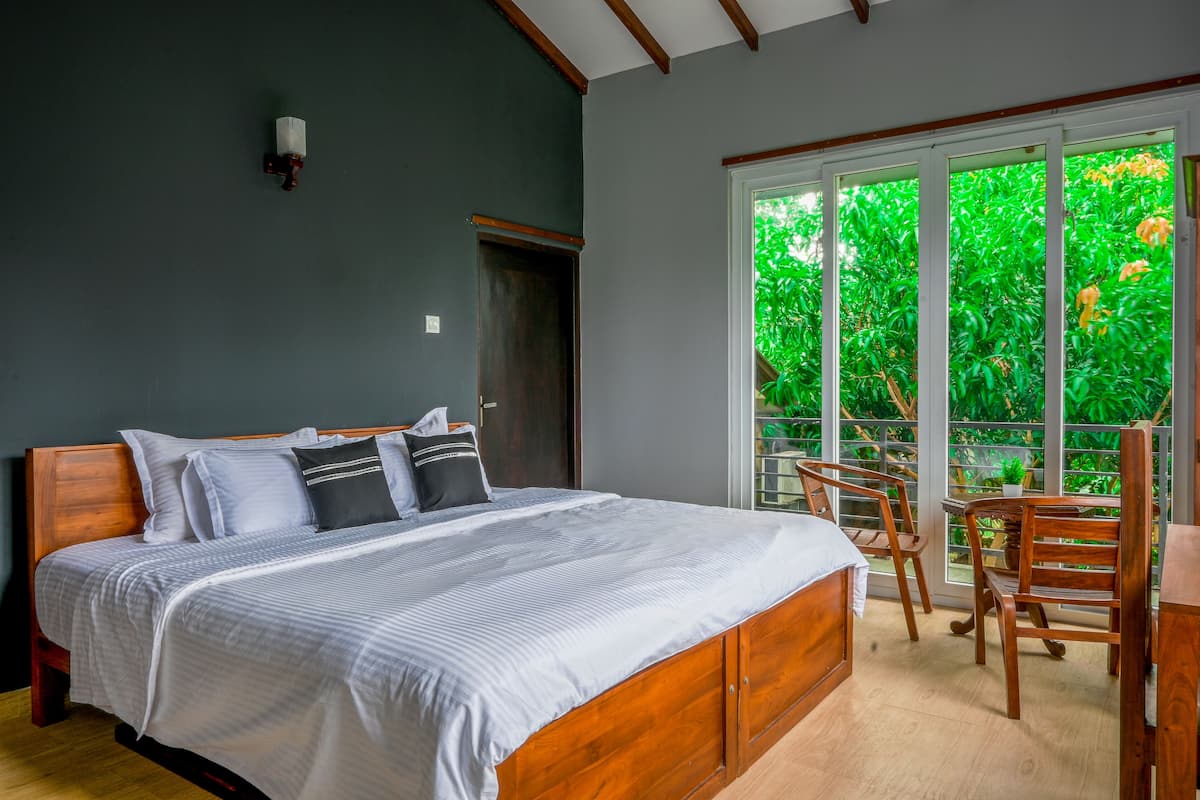 A bedroom with a large bed, a wooden headboard, a nightstand, and a window with a view of greenery.