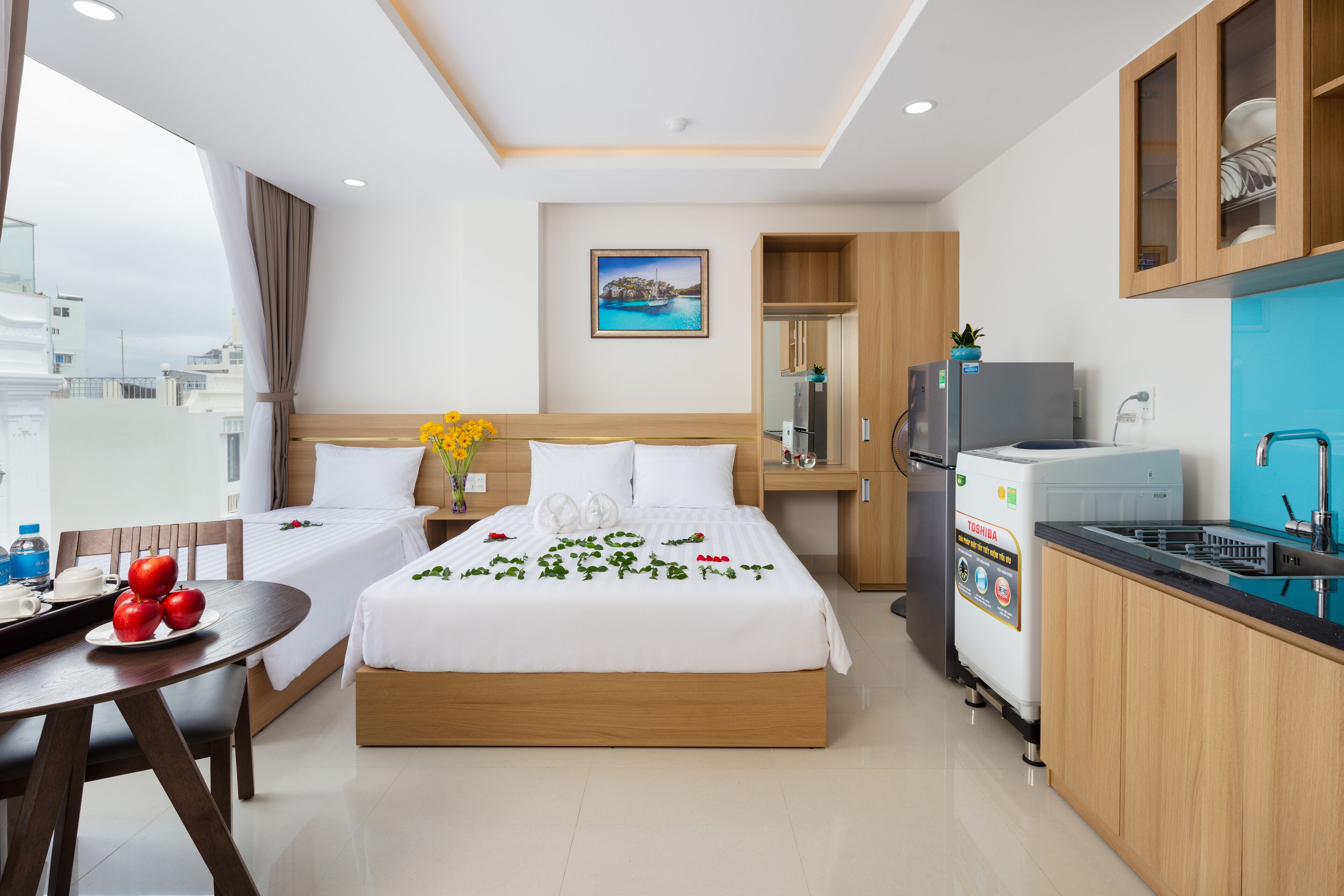 A hotel room with a bed, a small dining table with apples, a refrigerator, and a view of the city.