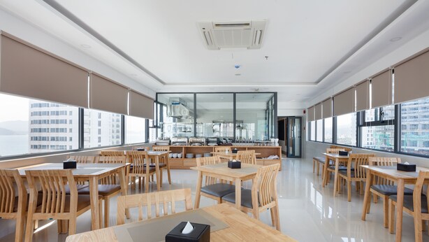 A modern dining area with wooden tables and chairs, large windows, and a view of the city. - XO Hotel & Apartments (Nha Trang)