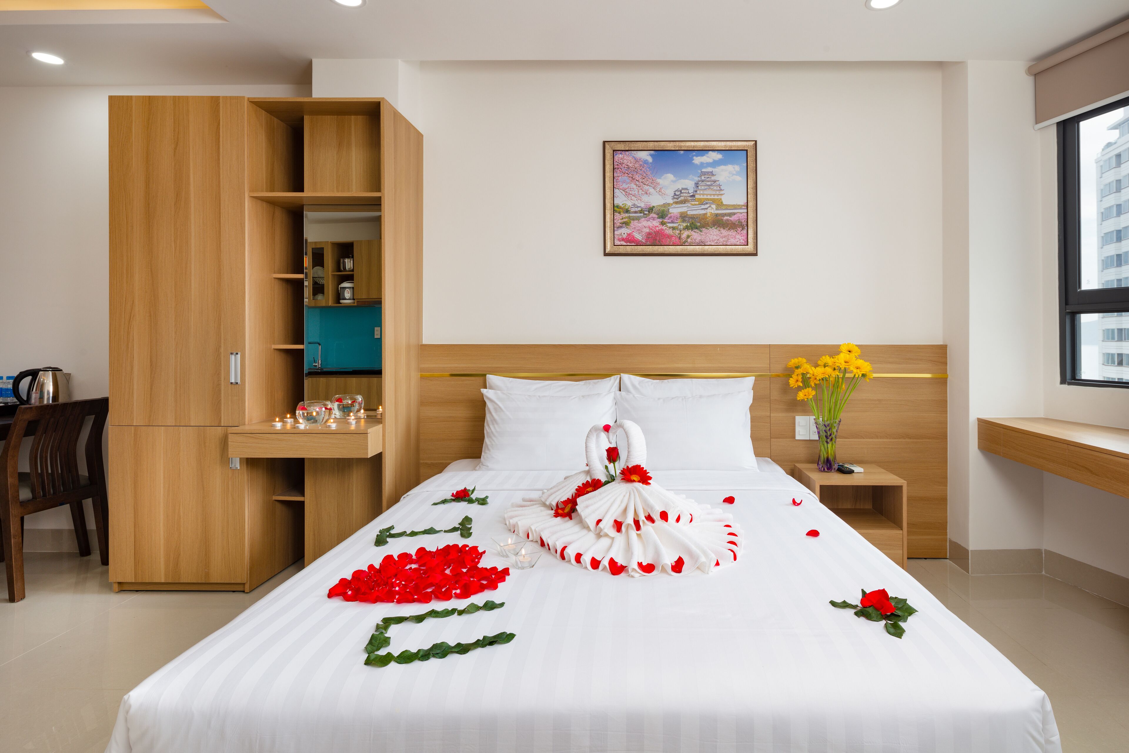 A hotel room with a bed, a wooden wardrobe, a small table with a vase of yellow flowers, and a painting on the wall.