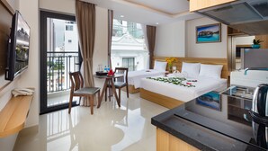 A hotel room with a bed, a desk, a chair, a small table, a TV, a kitchen area, and a balcony with a view of buildings. - XO Hotel & Apartments (Nha Trang)