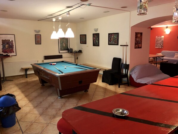 A pool table with balls, a red table, and a blue trash bin in a room with a tiled floor, a white radiator, and framed artwork on the walls. - Former Pub 3 for Stag do men's cave bachelor, bachelorette party for sleepers (IX. kerület)