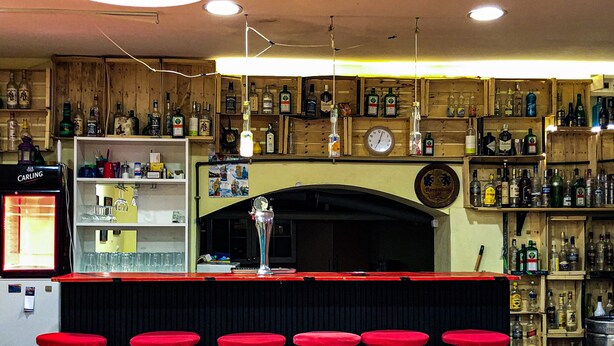 A bar with wooden shelves stocked with liquor bottles, a clock, and red bar stools. - Former Pub 3 for Stag do  men's cave  bachelor, bachelorette party for sleepers (IX. kerület)