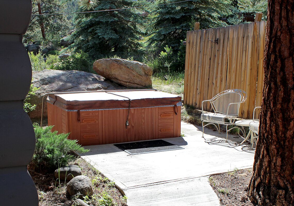 A hot tub with a wooden exterior, a patio area with outdoor furniture, and a wooden fence in a natural setting.