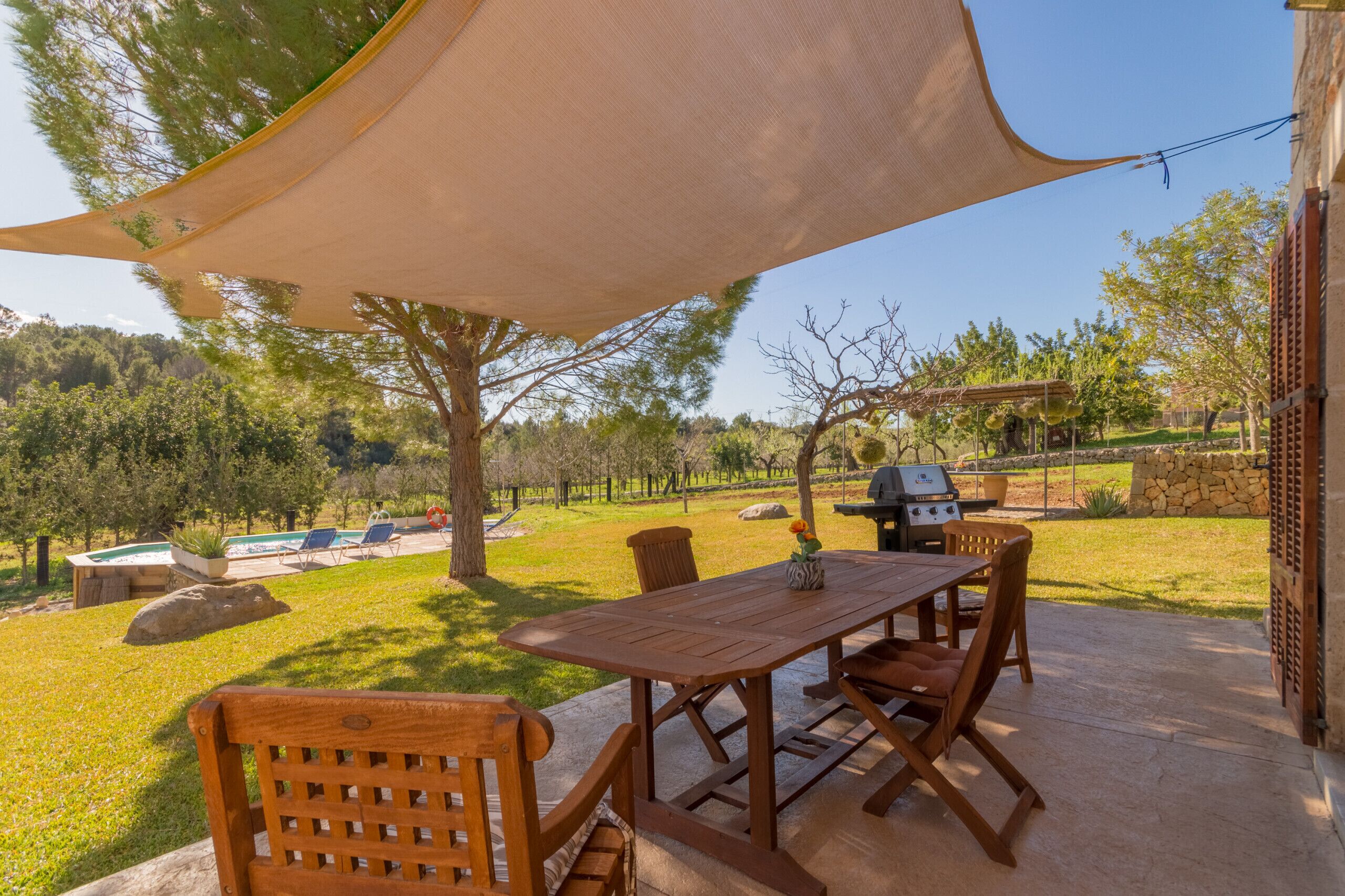 An outdoor patio with a wooden table and chairs, a barbecue grill, and a swimming pool in the background.