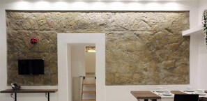 Ein Zimmer mit einer Steinwand, einem an der Wand befestigten Flachbildfernseher, einem Holztisch mit einer Schüssel und einem Durchgang zu einem anderen Raum.