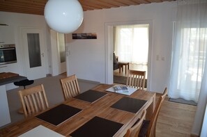 A dining area with a wooden table and chairs, a kitchen with an oven, and a view of a dining table through a doorway.