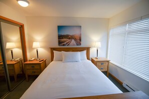 A hotel room with a large bed, two bedside tables with lamps, a wooden headboard, a painting above the bed, a mirror on one wall, and a window with blinds.