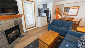 A living room with a fireplace, a blue sofa, a wooden coffee table, and a kitchen in the background.